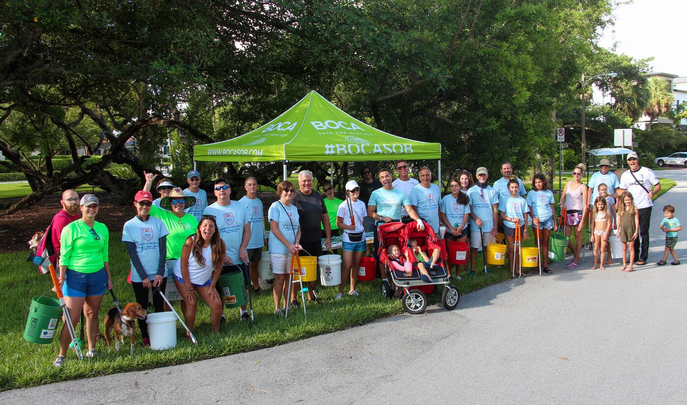 A large group of neighbors of all ages gathered on the grass with buckets, grabbers, and a canopy tent before a community beach cleanup.