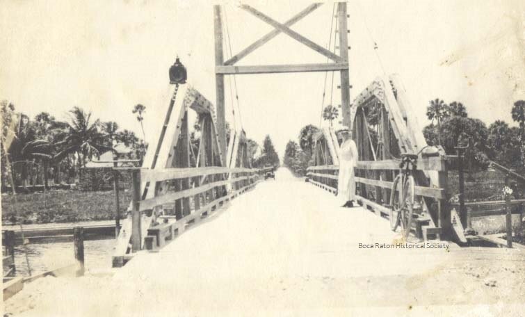 A sepia photograph of the original wooden Palmetto Park Road swing bridge, with a man in white standing beside a bicycle on the plank deck and palms lining the banks.